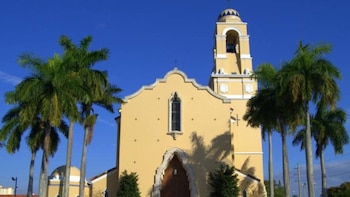 Fachada de una iglesia amarilla con campanario y ventanas arqueadas, rodeada de palmeras de troncos altos y esbeltos bajo un cielo azul despejado
