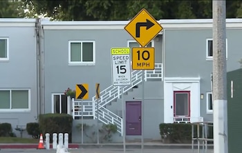Varias señales de tráfico, incluyendo una de límite de velocidad escolar de 15 mph y otra de 10 mph, frente a un edificio gris con escaleras blancas