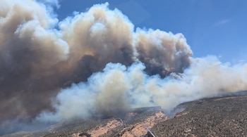 Gran columna de humo blanco y gris elevándose sobre montañas rocosas y un cañón con vegetación dispersa bajo un cielo azul parcial