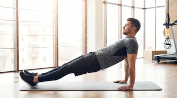 Un hombre está realizando una plancha invertida sobre una colchoneta gris en un gimnasio con ventanales y suelo de madera.