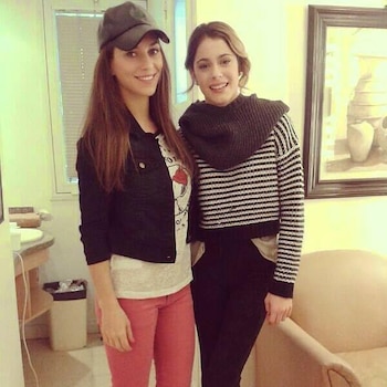 Dos mujeres de pie en un interior; una viste chaqueta y gorra oscura, y la otra lleva un suéter a rayas horizontales