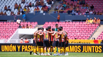 Grupo de futbolistas con uniforme de franjas rojas y amarillas abrazados en el campo frente a la tribuna del estadio.