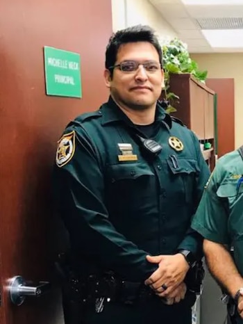 Un hombre con uniforme de policía verde, gafas de marco oscuro y placa dorada permanece de pie en una oficina junto a una puerta