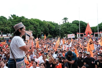 Keiko Fujimori, con camiseta blanca y un tocado de colores, habla en un micrófono frente a una gran multitud. La gente lleva camisetas y banderas naranjas, con árboles y un monumento al fondo