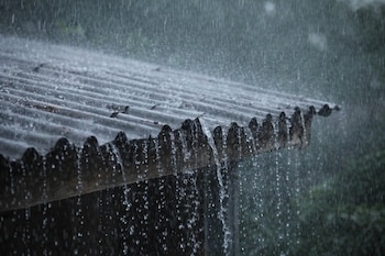 Lluvia moderada cayendo sobre un techo de lámina corrugada gris oscuro, con el agua goteando visiblemente desde el borde y salpicando.