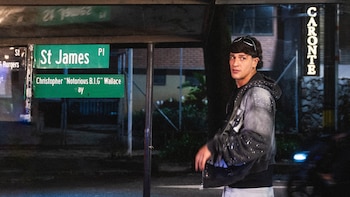 Un joven con cabello oscuro y gafas de sol en la cabeza, vistiendo una chaqueta gris con detalles blancos, mira hacia la izquierda junto a letreros callejeros