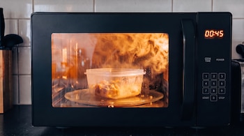 Horno de microondas negro con temporizador digital encendido y fuego con humo alrededor de un envase de plástico en su interior.