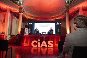 Tres hombres participan en una presentación sobre un escenario iluminado de rojo con letras gigantes del CIAS y una pantalla de fondo