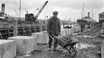 Fotografía en blanco y negro de un hombre con gorra que sostiene una carretilla con cuerdas entre bloques de hormigón y barcos al fondo.
