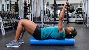 Mujer con camiseta turquesa tumbada sobre un rodillo azul en un gimnasio, levantando una pesa rusa negra. Brazo extendido. Equipos de fondo
