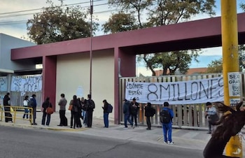 Grupo de personas frente a la entrada de un edificio universitario con dos pancartas que dicen 'LA TOMA DEL LOCAL' y '¿DONDE ESTÁN 8 MILLONES?'
