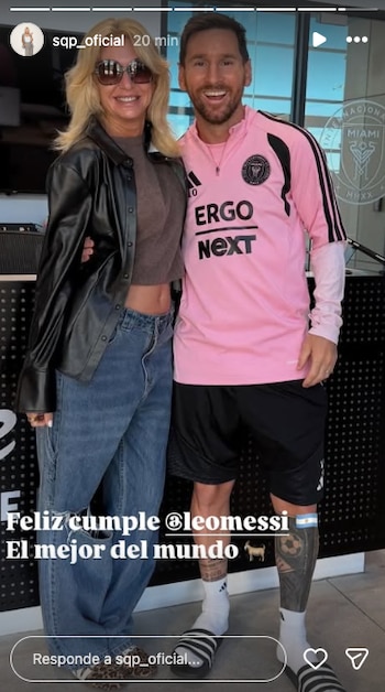 Lionel Messi sonríe junto a una mujer rubia con gafas de sol. Messi viste camiseta rosa, shorts negros, sandalias y muestra tatuajes en sus piernas