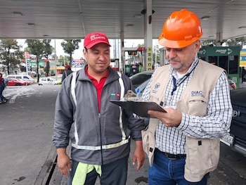 Dos hombres en una gasolinera. Uno con casco naranja y chaleco beige, el otro con gorra roja y chaqueta gris. El de casco revisa un portapapeles