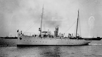 Fotografía en blanco y negro de un buque naval con dos mástiles, chimenea humeante y varias personas en cubierta, navegando en aguas tranquilas