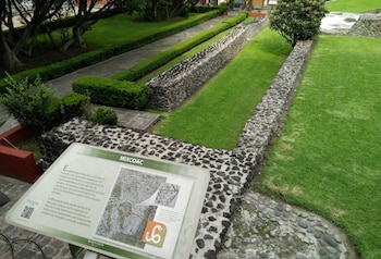 Cartel informativo con texto y mapa de sitio arqueológico Mixcoac, muros de piedra, césped verde, arbustos recortados y árboles en un parque