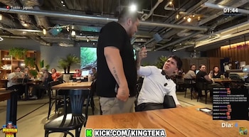 Dos hombres en un restaurante. Uno de pie, corpulento, con camiseta negra. El otro sentado, con camisa blanca y corbata de lazo, reacciona. Mesas y comensales al fondo