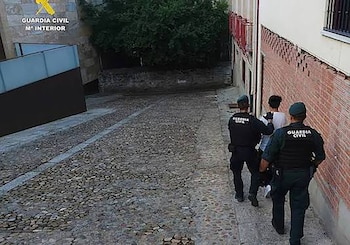 Dos agentes de la Guardia Civil uniformados caminan de espaldas junto a una persona con camiseta blanca por una calle empedrada