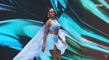 Una mujer con traje azul brillante de dos piezas y una capa clara desfila sobre un escenario iluminado