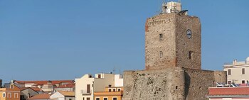 Vista frontal de una torre de piedra gris y marrón con un reloj y varias ventanas pequeñas, sobresaliendo de edificios con tejados naranjas bajo un cielo azul claro