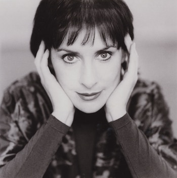 Retrato en blanco y negro de Enya mirando directamente a la cámara. Tiene el cabello oscuro con flequillo y sus manos cubren los lados de su rostro