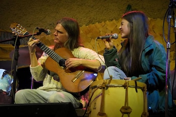 Una persona con cabello largo toca una guitarra acústica con los ojos cerrados frente a un micrófono en un escenario iluminado