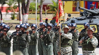 Grupo de soldados de uniforme verde y camuflado, con cascos y rifles, en formación. Un militar sostiene una bandera roja. Un tanque militar se observa al fondo