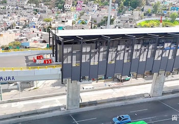 Estación elevada con paneles oscuros sobre un viaducto de concreto, con vehículos en la avenida inferior y casas sobre una colina