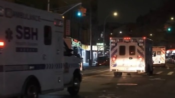 Vista nocturna de dos ambulancias blancas con luces rojas intermitentes en una calle oscura de la ciudad, con farolas, semáforos y edificios iluminados