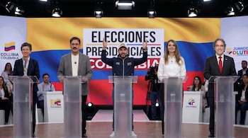 Cinco candidatos presidenciales de pie en un escenario de debate con podios transparentes. Un letrero grande y una bandera colombiana se ven al fondo.