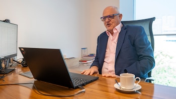Hombre con anteojos y chaqueta azul sentado en un escritorio de madera con una computadora portátil, un monitor y una taza de café