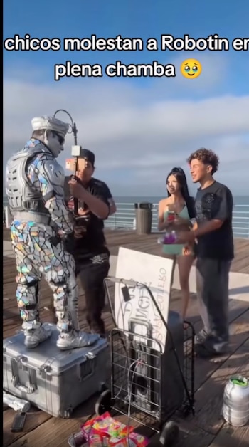 Hombre vestido de robot en muelle junto a tres jóvenes, uno filma con teléfono. Hay un carrito con equipos y un letrero. Cielo azul y océano
