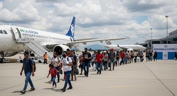 Grupo de personas camina en fila sobre la pista de un aeropuerto. Se observa un avión blanco, escaleras de embarque y un cartel de 'Bienvenida a Guatemala'.