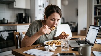 Mujer sentada a una mesa comiendo con tenedor y sosteniendo una porción de pizza junto a una computadora portátil.