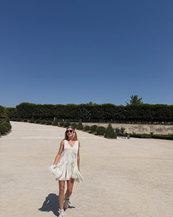 Una mujer con vestido blanco, pañuelo en la cabeza y gafas de sol camina por un sendero de grava en un parque con setos verdes