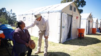 Hombre inclinado con gorra y camisa blanca conversa con mujer sentada en silla, delante de varias tiendas de campaña blancas sobre césped con árboles al fondo