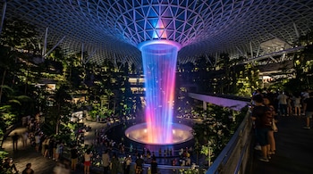 Una gran cascada interior iluminada con luces púrpuras y azules bajo una cúpula de vidrio y metal, rodeada de plantas y personas.