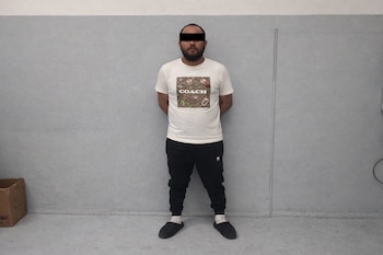Hombre barbudo con banda negra en los ojos, camiseta blanca, pantalón negro, calcetines blancos y zapatillas negras de pie frente a una pared gris.