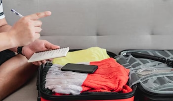 Manos sosteniendo un cuaderno y un bolígrafo sobre una maleta roja abierta con ropa doblada, un teléfono móvil negro y un accesorio de cámara