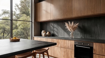Cocina moderna con gabinetes de madera clara, encimeras y mesa oscuras, un gran ventanal con vista a árboles, un horno y elementos decorativos como jarrones y una cesta con limones.