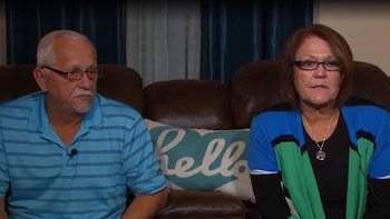 Un hombre y una mujer maduros con anteojos están sentados en un sofá de cuero frente a un cojín con la palabra hello