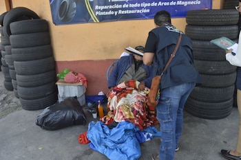 Una persona sentada, cubierta con una manta de colores, conversa con otra persona que sostiene un portapapeles, junto a pilas de neumáticos, una bolsa de basura y otros objetos en el suelo