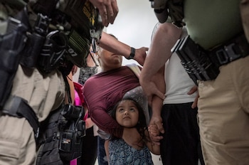 Un primer plano de un niño pequeño que mira hacia arriba mientras es sostenido por una mujer adulta, rodeados por las manos y torsos de varios adultos
