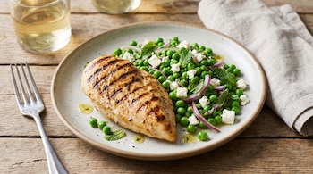 Pechuga de pollo grillé dorada en un plato, junto a una ensalada de arvejas verdes, cebolla morada, queso feta y hojas de menta, sobre una mesa de madera.