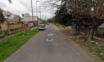 Calle asfaltada con un automóvil gris estacionado, casas bajas, postes del alumbrado público y árboles en la acera