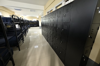 Dormitorio con literas de dos pisos y colchones azules. Fila de taquillas metálicas negras con cerraduras y ventilación. Piso de baldosas claras, paredes beige, ventanas