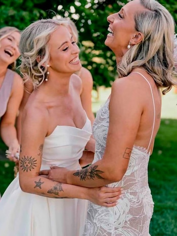 Dos mujeres rubias con vestidos de tonos claros se abrazan y ríen. Una tiene tatuajes en los brazos. Una tercera mujer rubia sonríe en el fondo