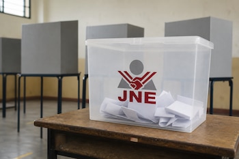 Una urna transparente con el logotipo del Jurado Nacional de Elecciones sobre una mesa de madera y cabinas de votación al fondo.