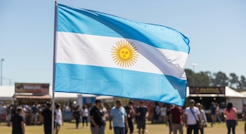 Bandera argentina flameando al aire libre, mostrando sus franjas celestes y blancas con el Sol de Mayo, bajo un cielo azul. Fondo desenfocado de un festival con gente.