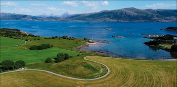 Vista aérea de un paisaje costero noruego con un amplio fiordo azul, montañas a lo lejos, campos verdes en primer plano y un camino serpenteante
