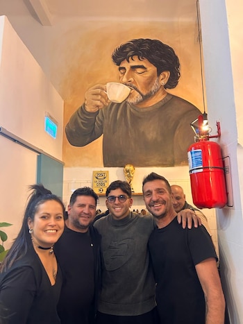 Cuatro personas posan sonrientes bajo un mural que representa a un hombre bebiendo café y un trofeo dorado en un estante